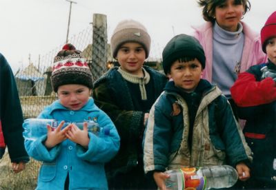Tschetschenische Kinder neben einem Zaun, alle mit Windjacke und Mütze. Einige halten eine Plastikflasche in der Hand.